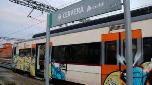 Un cartell de la parada de Cervera amb un tren a darrere (Anna Berga, ACN)